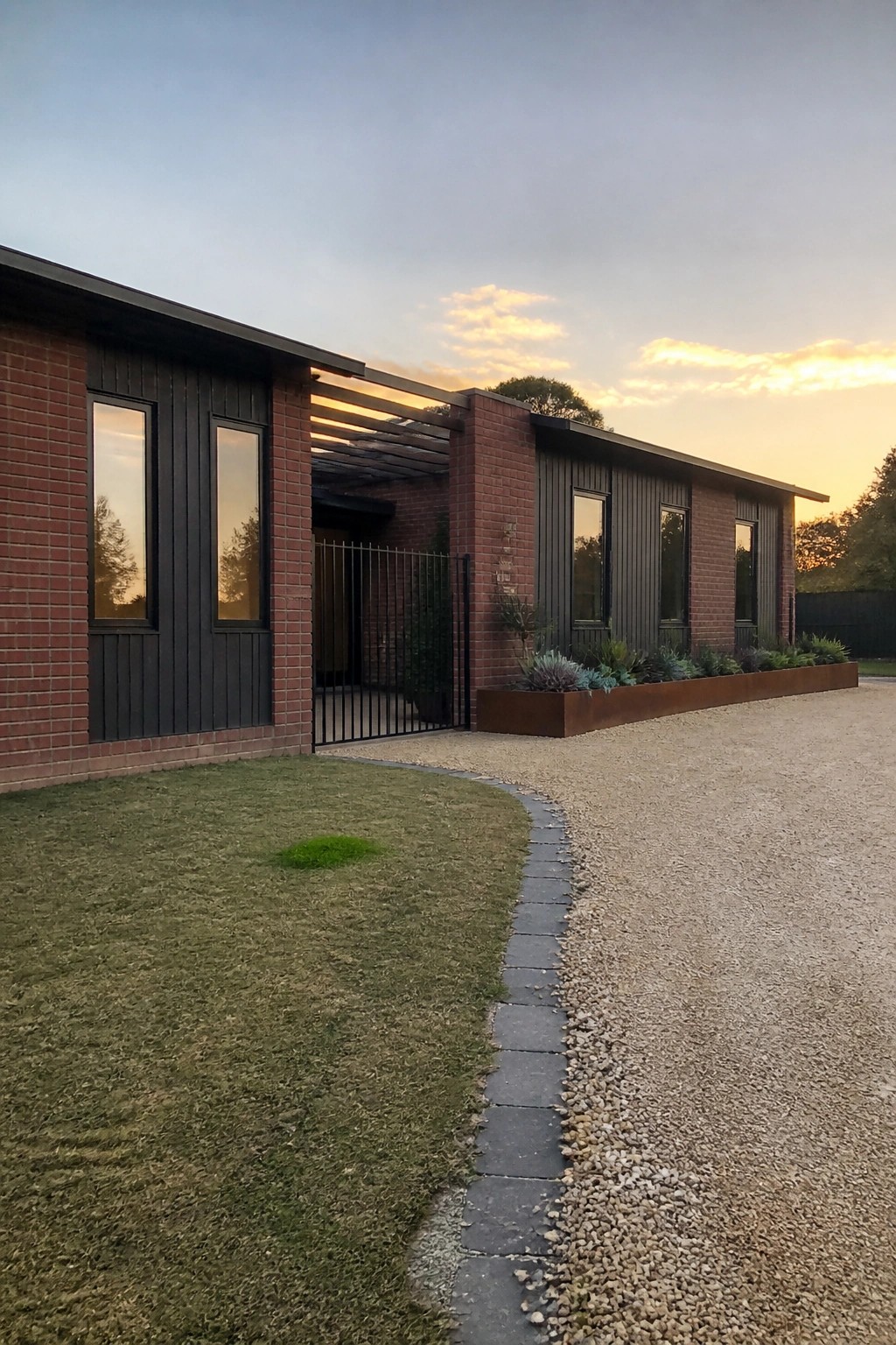 Modern brick home with dark panels, gravel driveway, metal planter at sunset