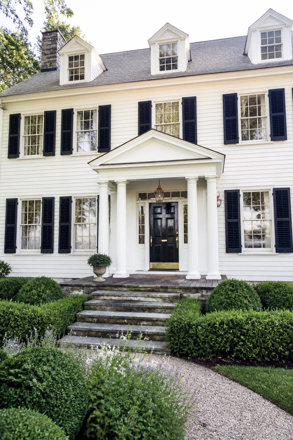 White colonial house with black shutters, columned portico, stone steps, and lush landscaping