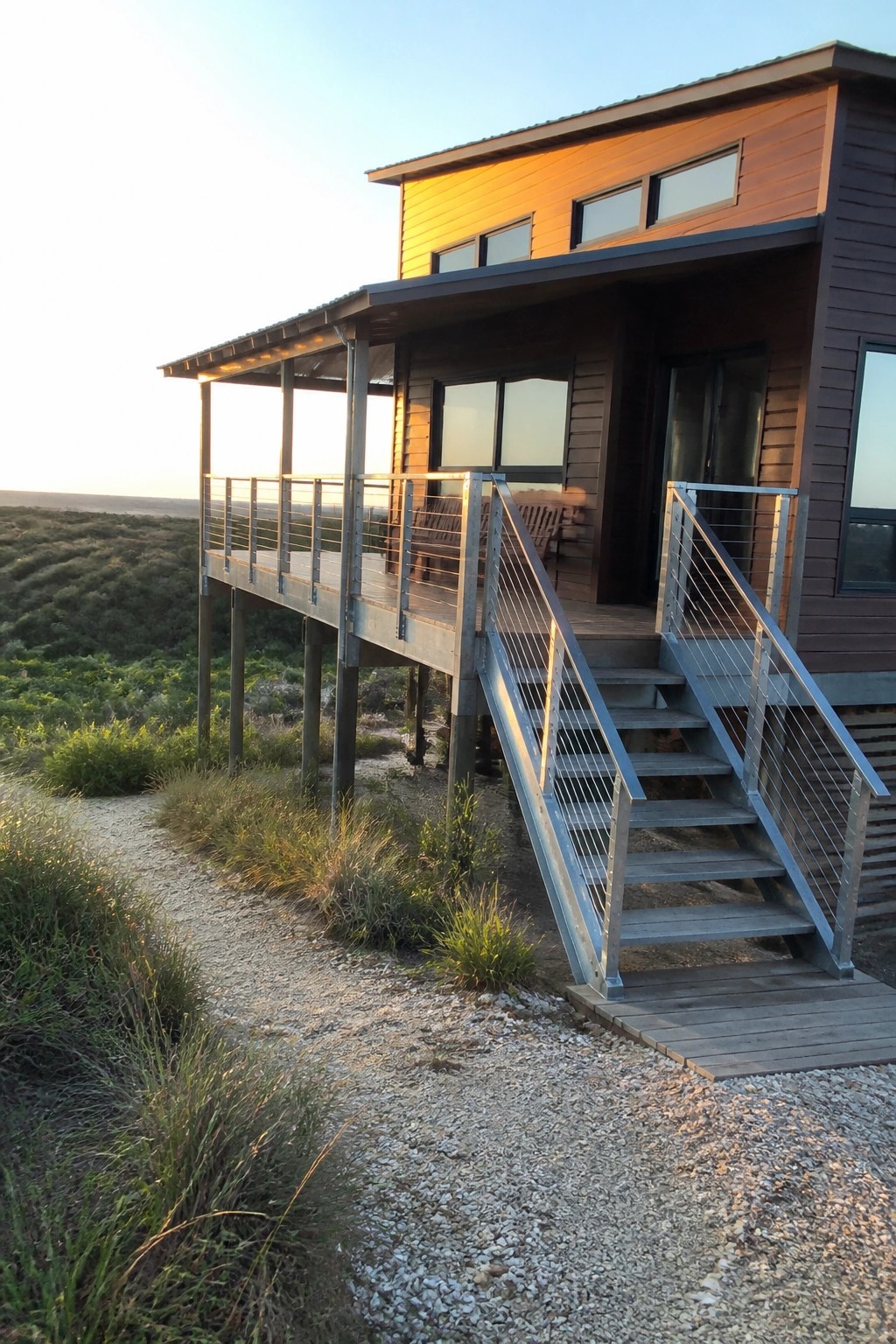 A modern two-story house with horizontal brown siding elevated on posts above a grassy dune, featuring a wooden deck and metal-railed stairs leading to a gravel path.