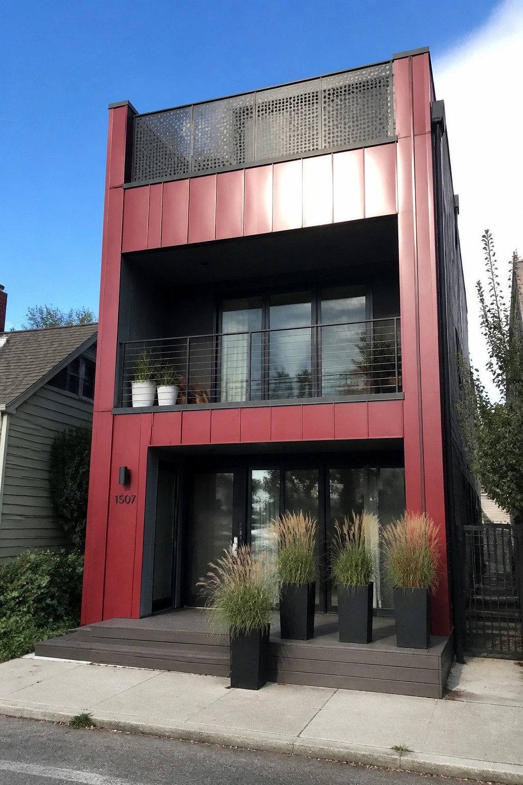 A modern three-story house with red metal siding, a second-floor balcony with railings, and tall potted grasses at the entrance.