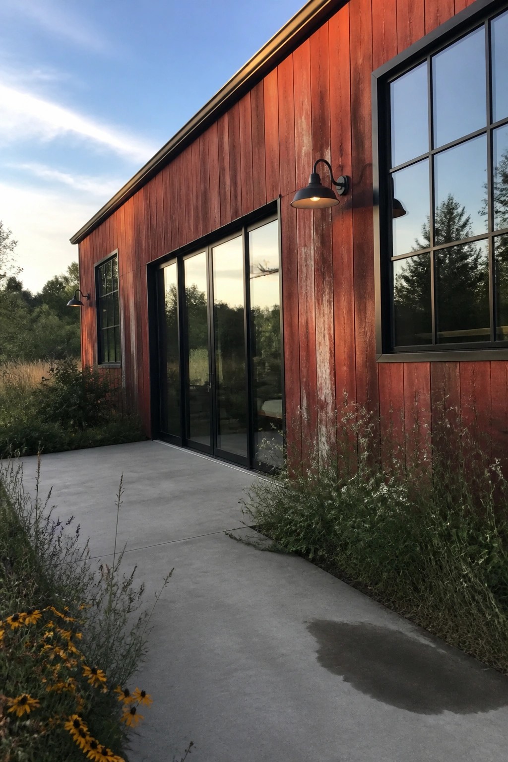 A modern house exterior with vertical red wood siding, black-framed windows, wall sconces, and a concrete walkway bordered by plants.
