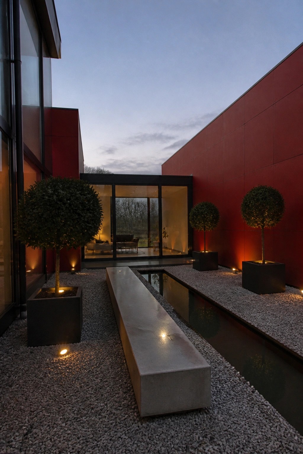 Modern courtyard at dusk with red walls, concrete bench, water feature, and lit topiary trees.