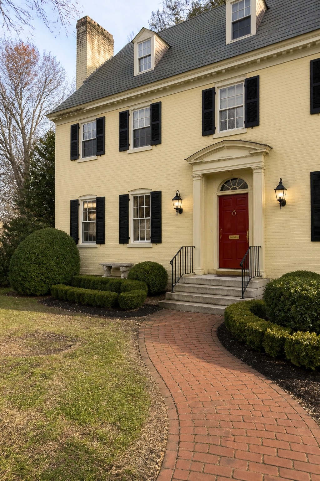 Yellow two-story brick house with red door, black shutters, and brick pathway.