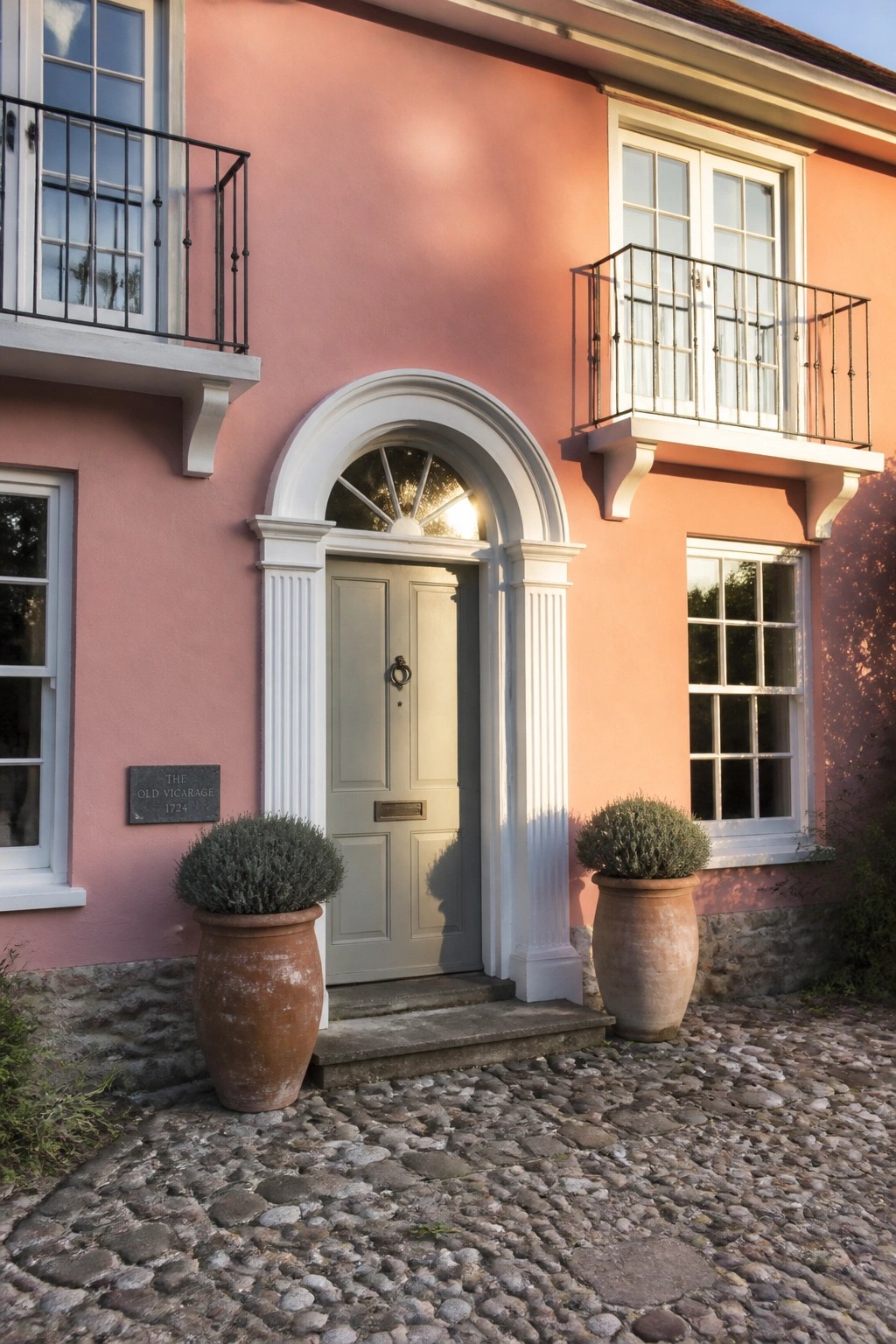 A pink cottage exterior featuring a sage green door beneath a white arched surround, with two large terracotta pots on a cobblestone path.