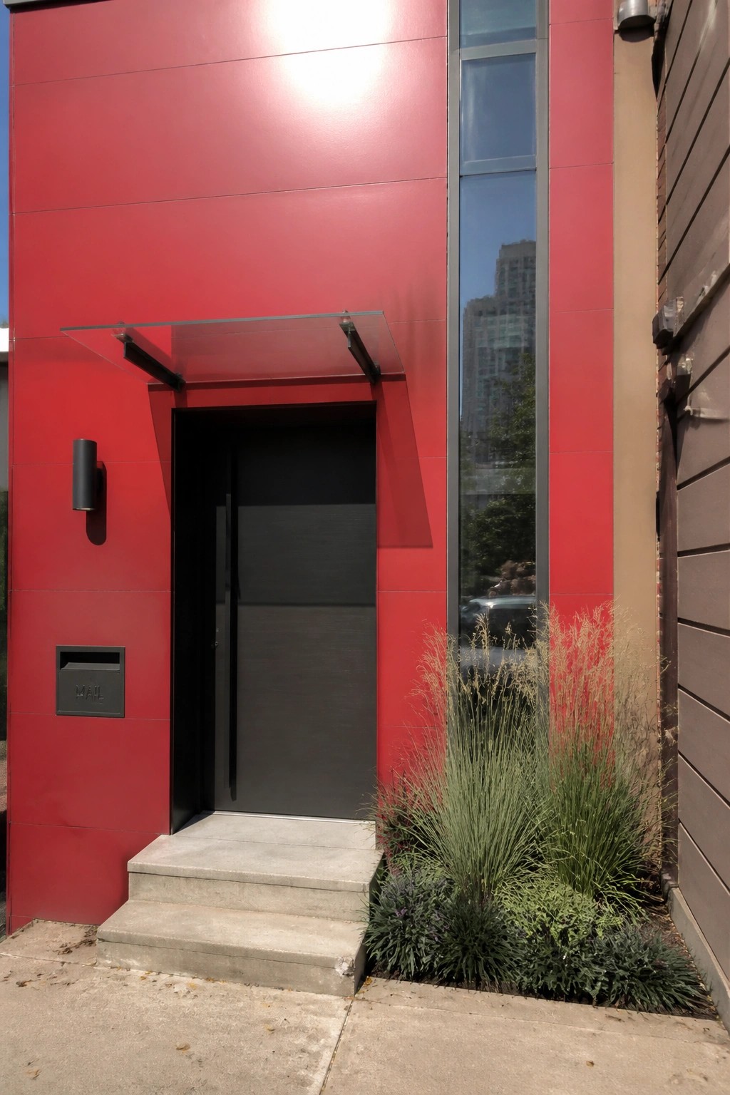 Red building entrance with black door, glass awning, concrete steps, and plants