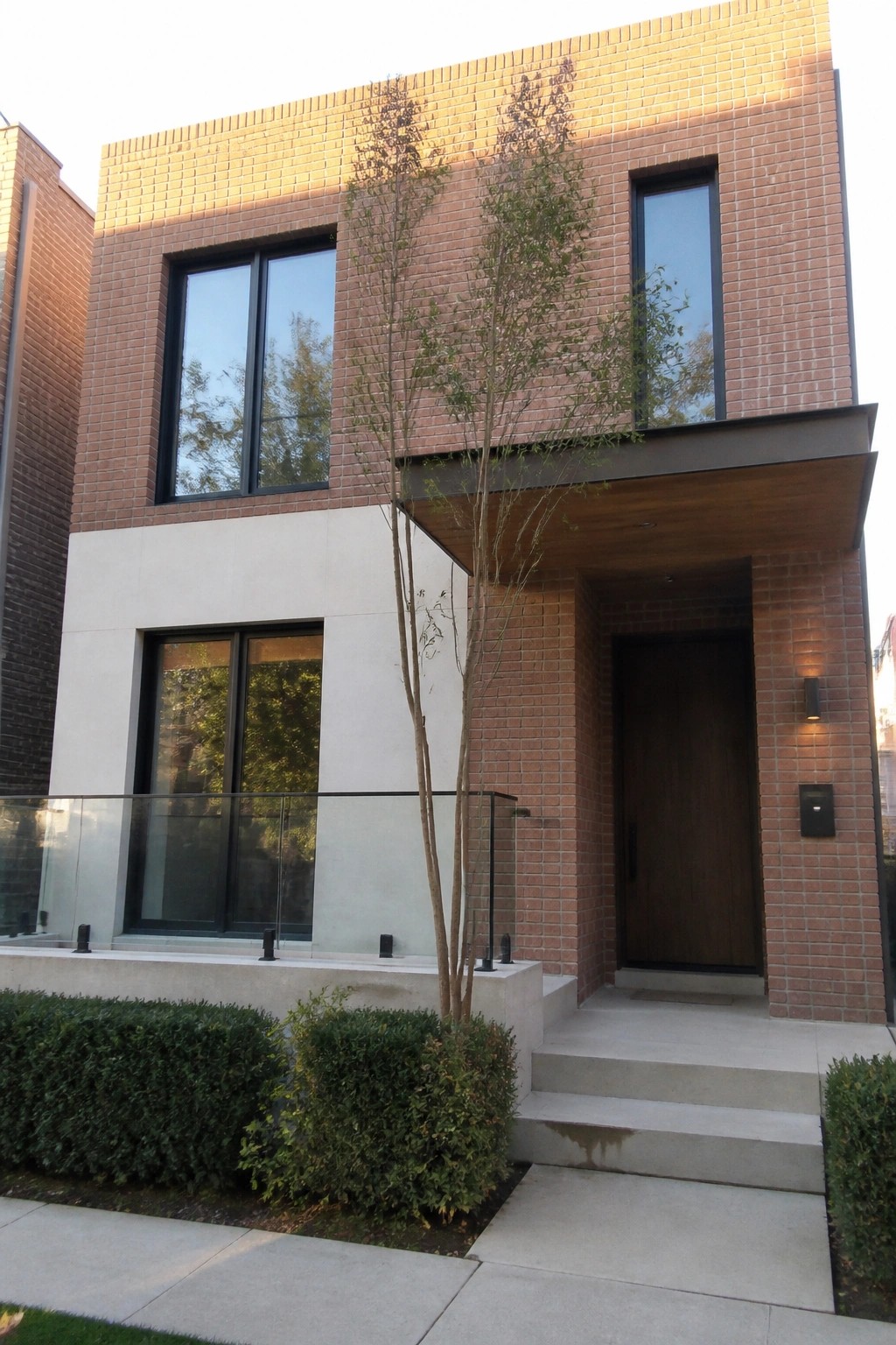 A modern two-story red brick house with large black-framed windows, light concrete panels on the lower facade, a dark wood front door under a wooden canopy, and concrete steps with glass railings.