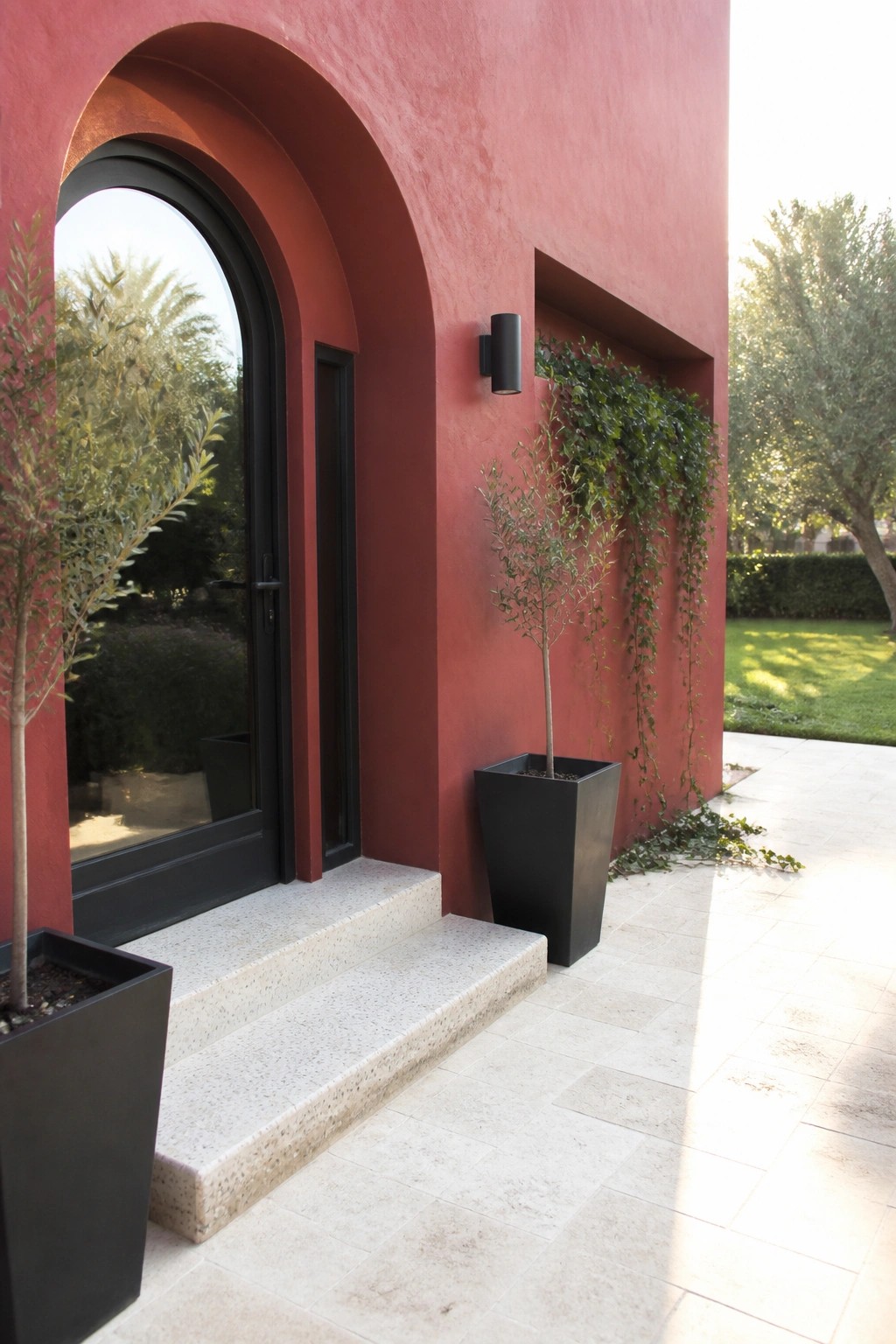 Red stucco house exterior with black arched door, stone steps, and black planters holding small trees.