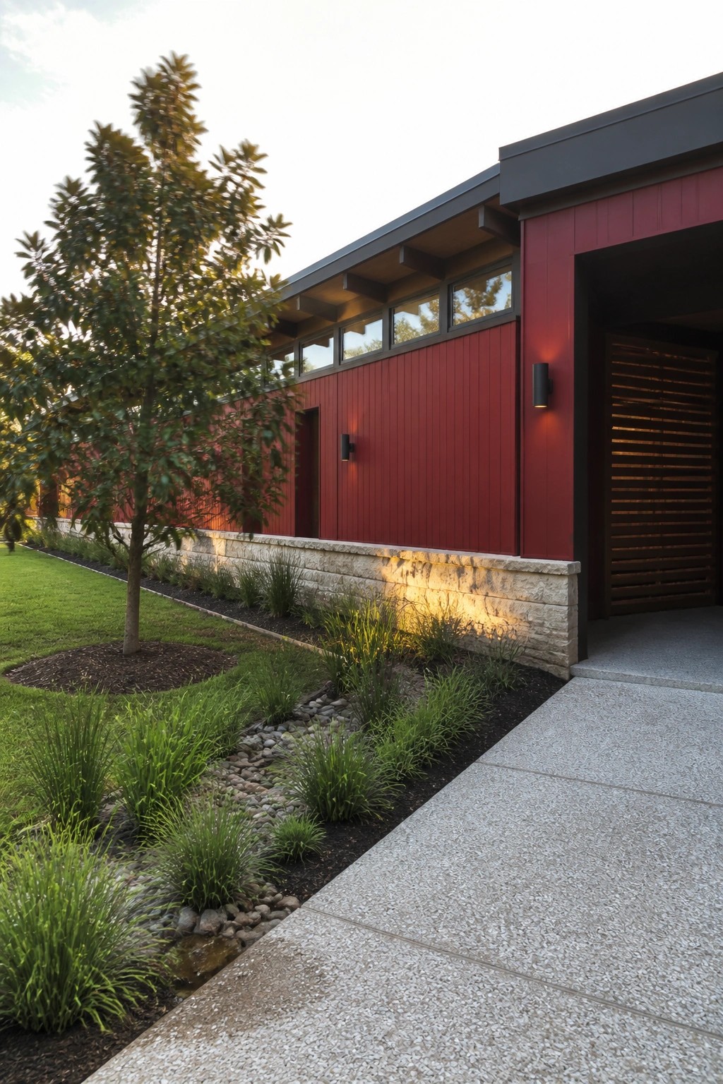 A modern red house with vertical siding, a light stone base, and a landscaped garden bed along the front next to a concrete driveway.