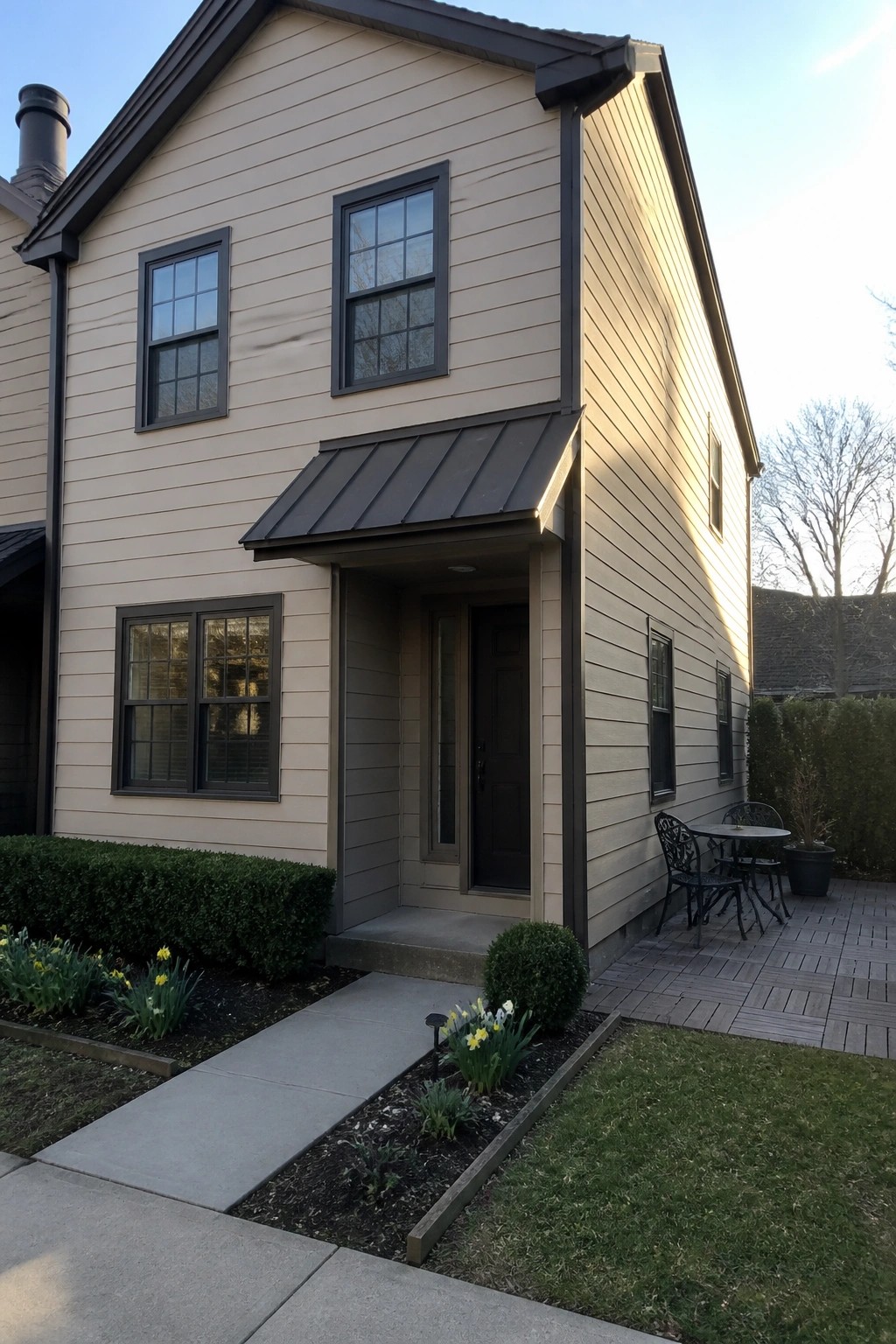 A two-story suburban house with beige siding, dark trim around the windows and door, and a small front patio area.