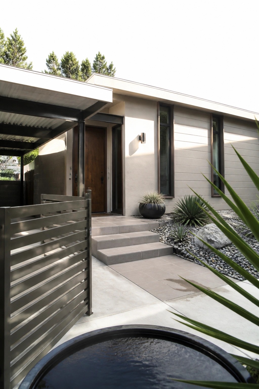 Modern suburban house with horizontal beige siding, dark-framed windows, wooden front door, concrete steps, and gravel landscaping with plants.