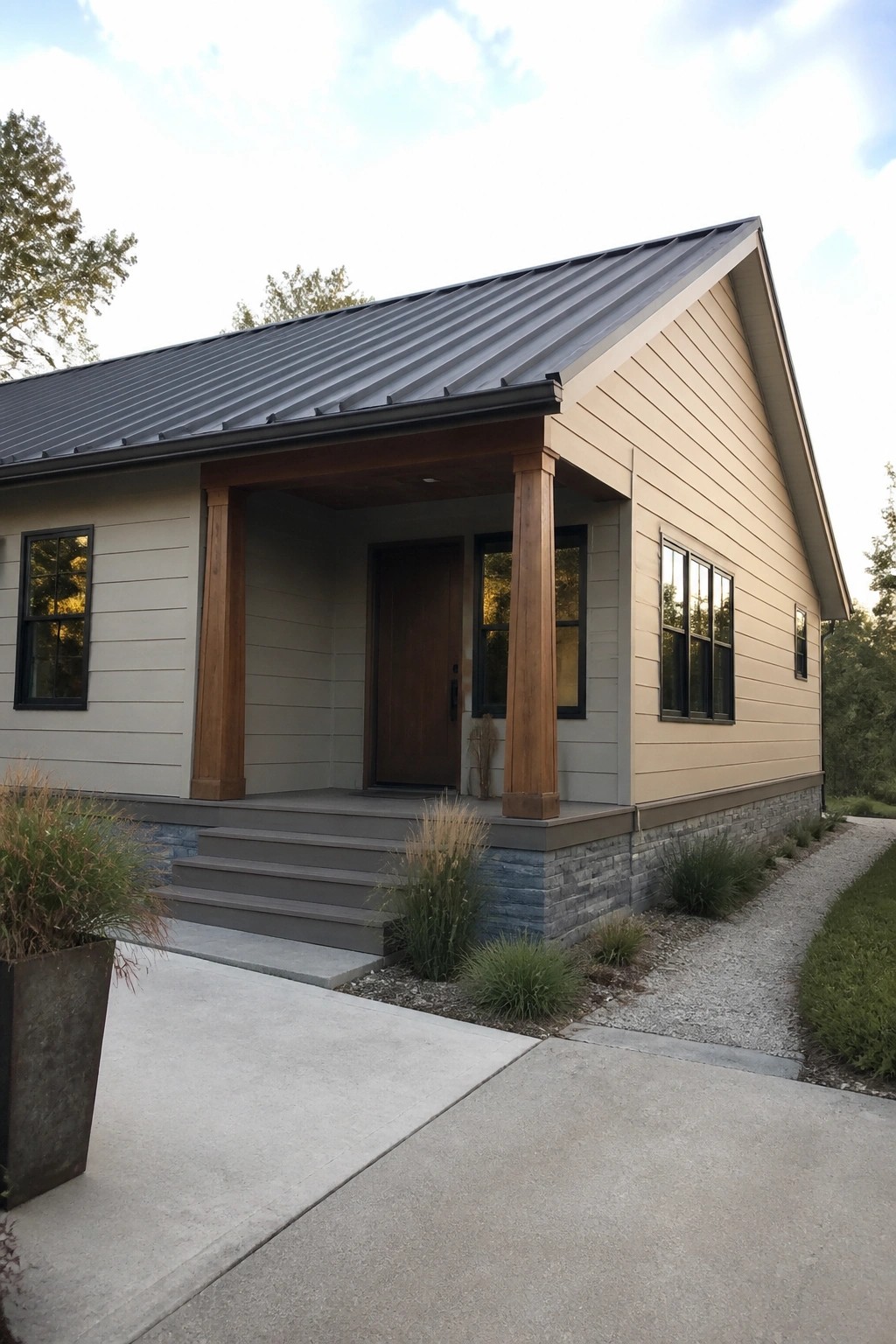 Front view of a single-story house with beige horizontal siding, a dark metal roof, wooden porch posts, and a stone foundation.