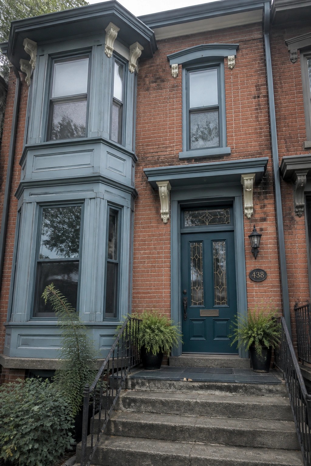Red brick townhouse with blue bay window, teal door, and fern-lined steps.
