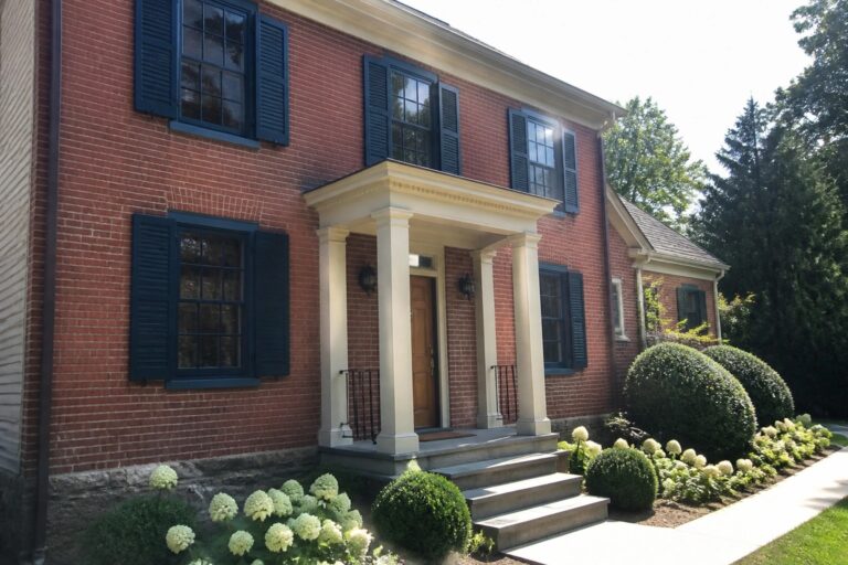 Red brick house with white columns, blue shutters, wooden door, and flower garden path