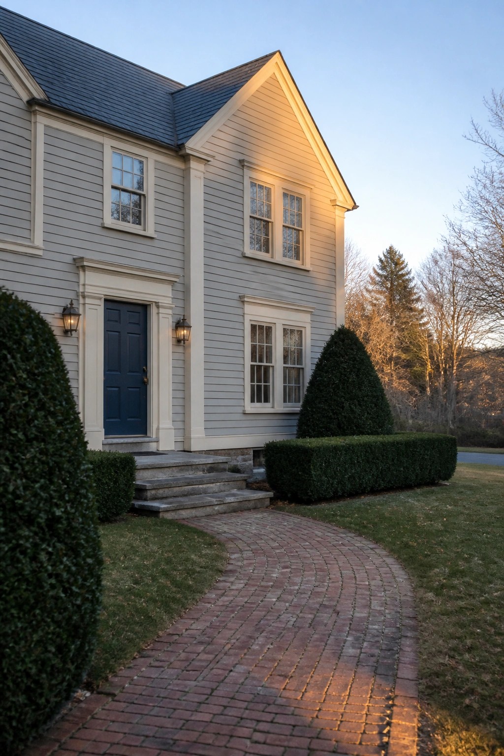 Gray house with blue door, curving brick path, and manicured hedges at dusk