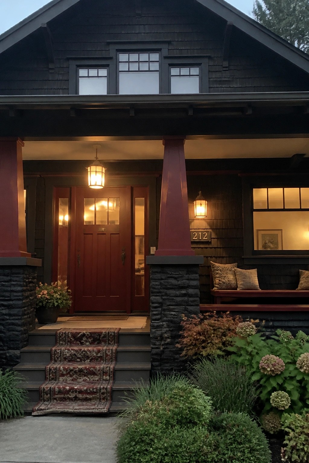 Dark shingled house with red door, lit lanterns, stone pillars, and flower-lined steps.
