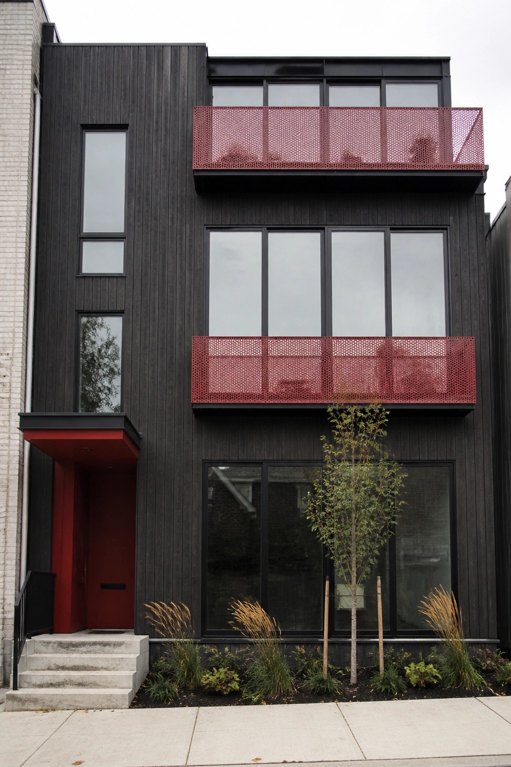 A modern two-story house with black wood siding, large windows, red perforated metal balconies on the upper levels, a red front door with a matching overhang, and small plants along the sidewalk.