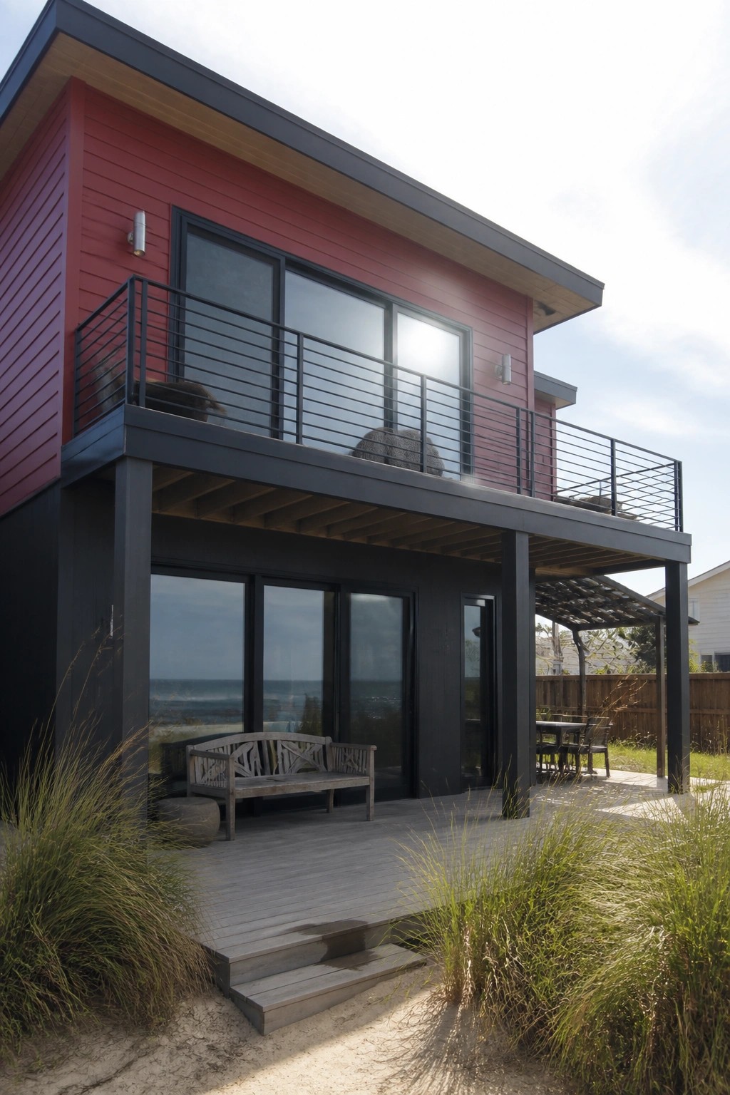 A two-story modern house with red upper siding, black lower structure and framing, large windows, and a deck overlooking the ocean.
