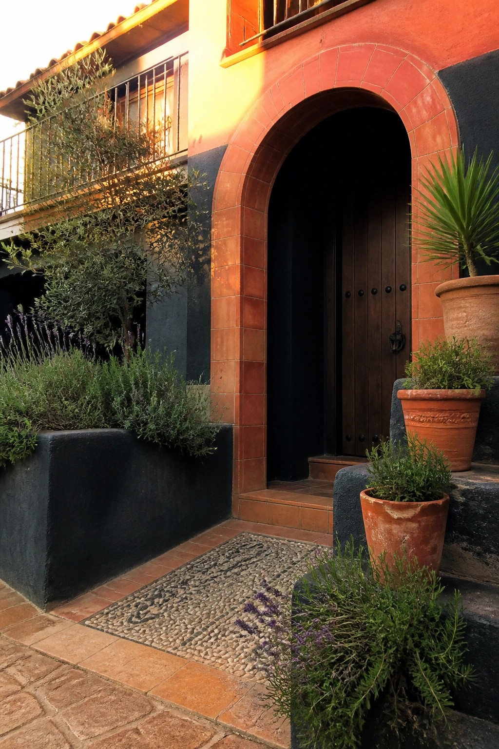 Red arched doorway with wooden doors, terracotta pots, plants, and pebble mosaic mat.