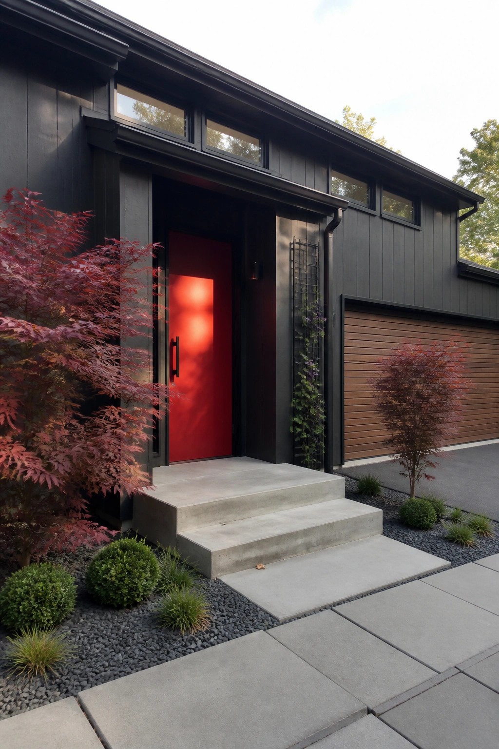 Modern house exterior with black siding, a bright red front door, concrete steps, and red foliage plants beside the entry.
