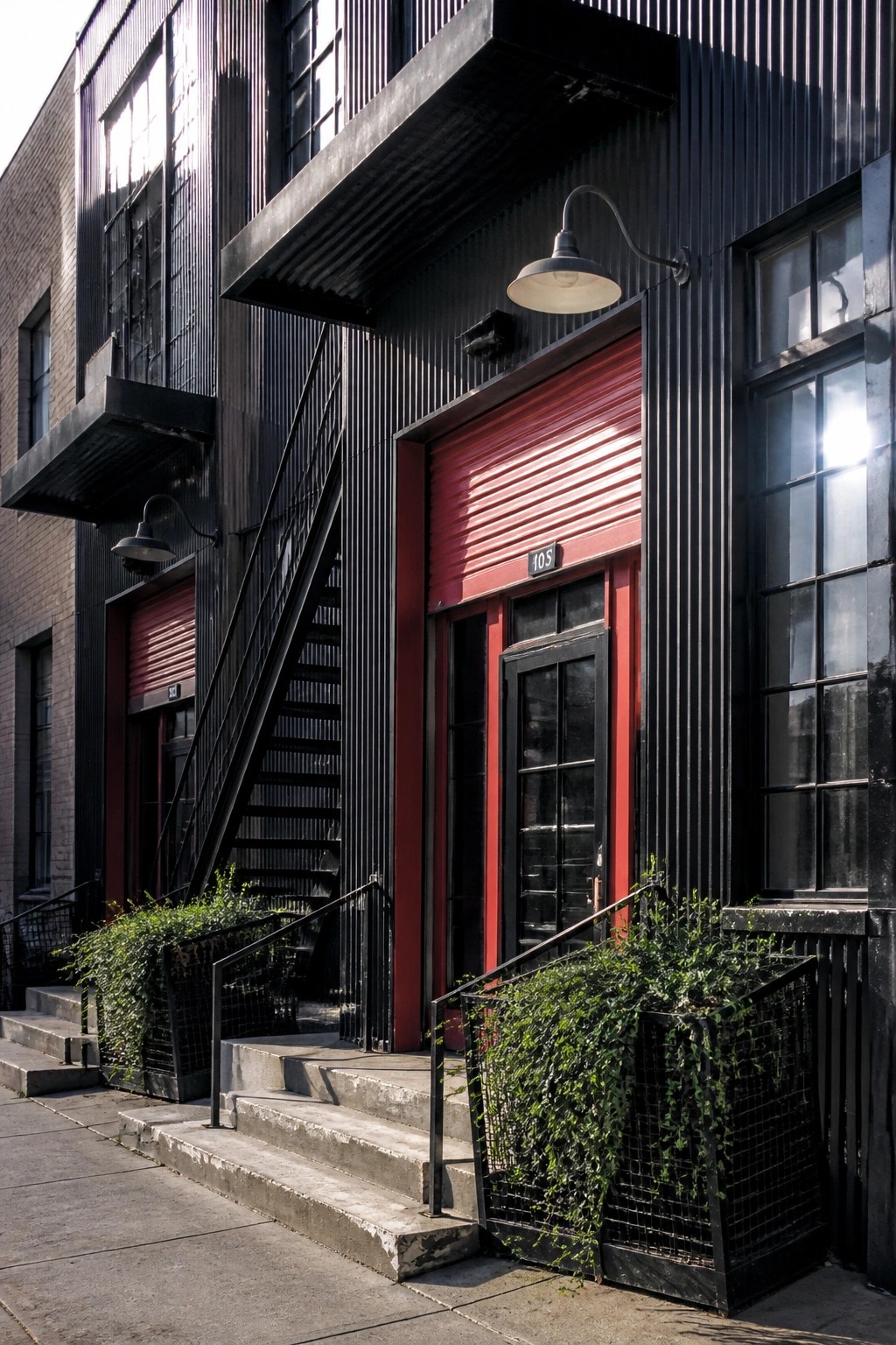 Black industrial building with red doors, fire escape stairs, and green planters.