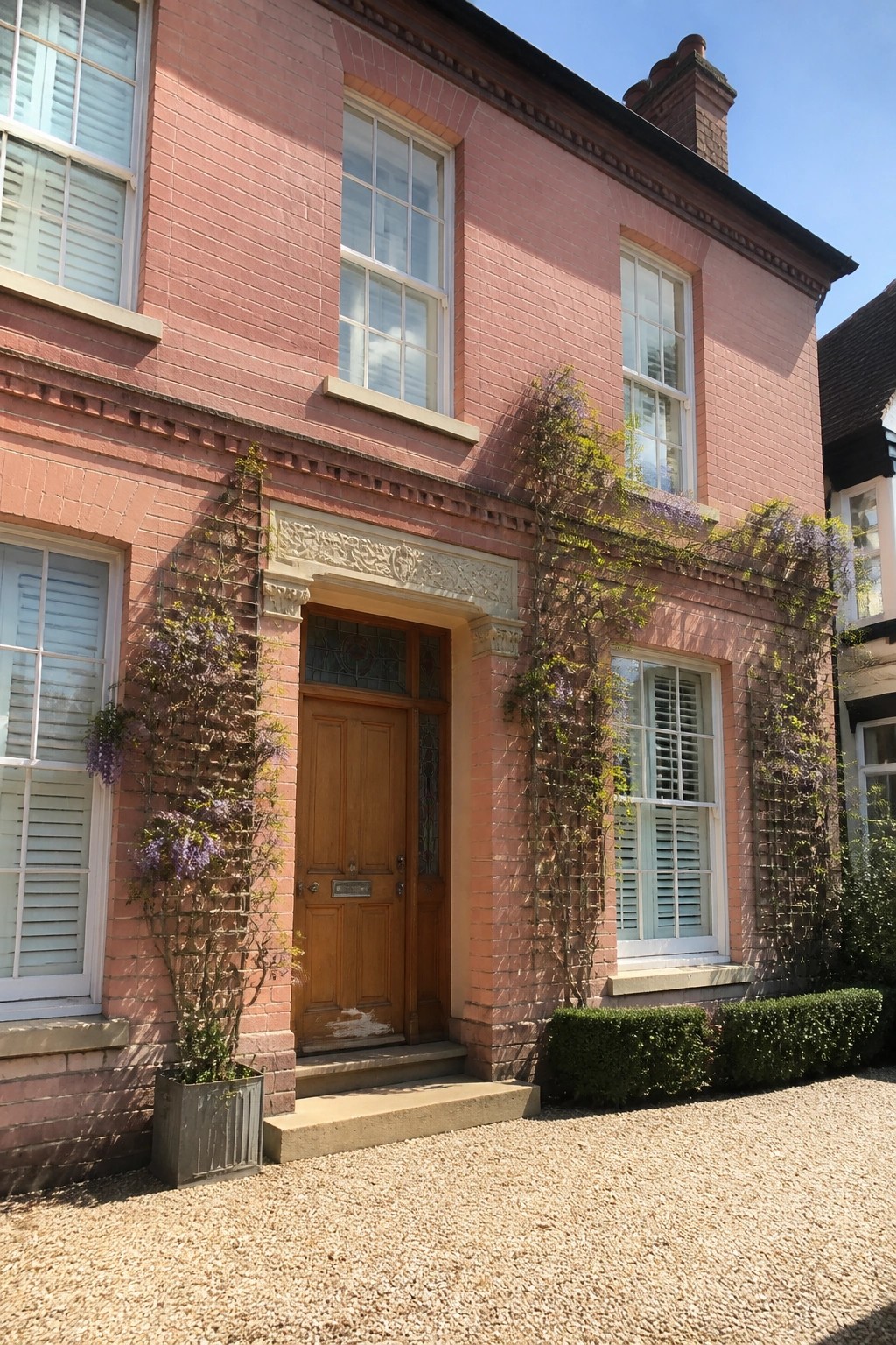 A two-story pink brick house with a wooden front door, carved stone surround, and climbing vines growing on trellises beside the entrance.