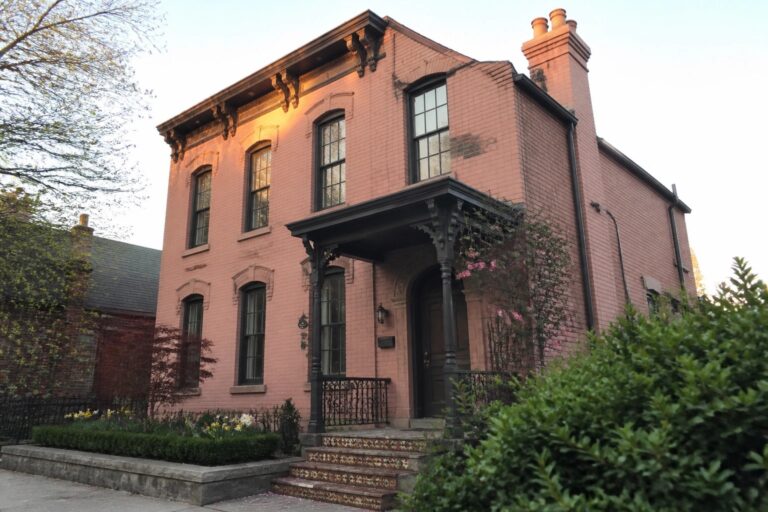 22 Textured Pink Brick House Exteriors That Preserve Historic Character A two-story pink brick house with an arched dark wood door, decorative porch columns, patterned tile stairs, and multiple windows with dark trim.