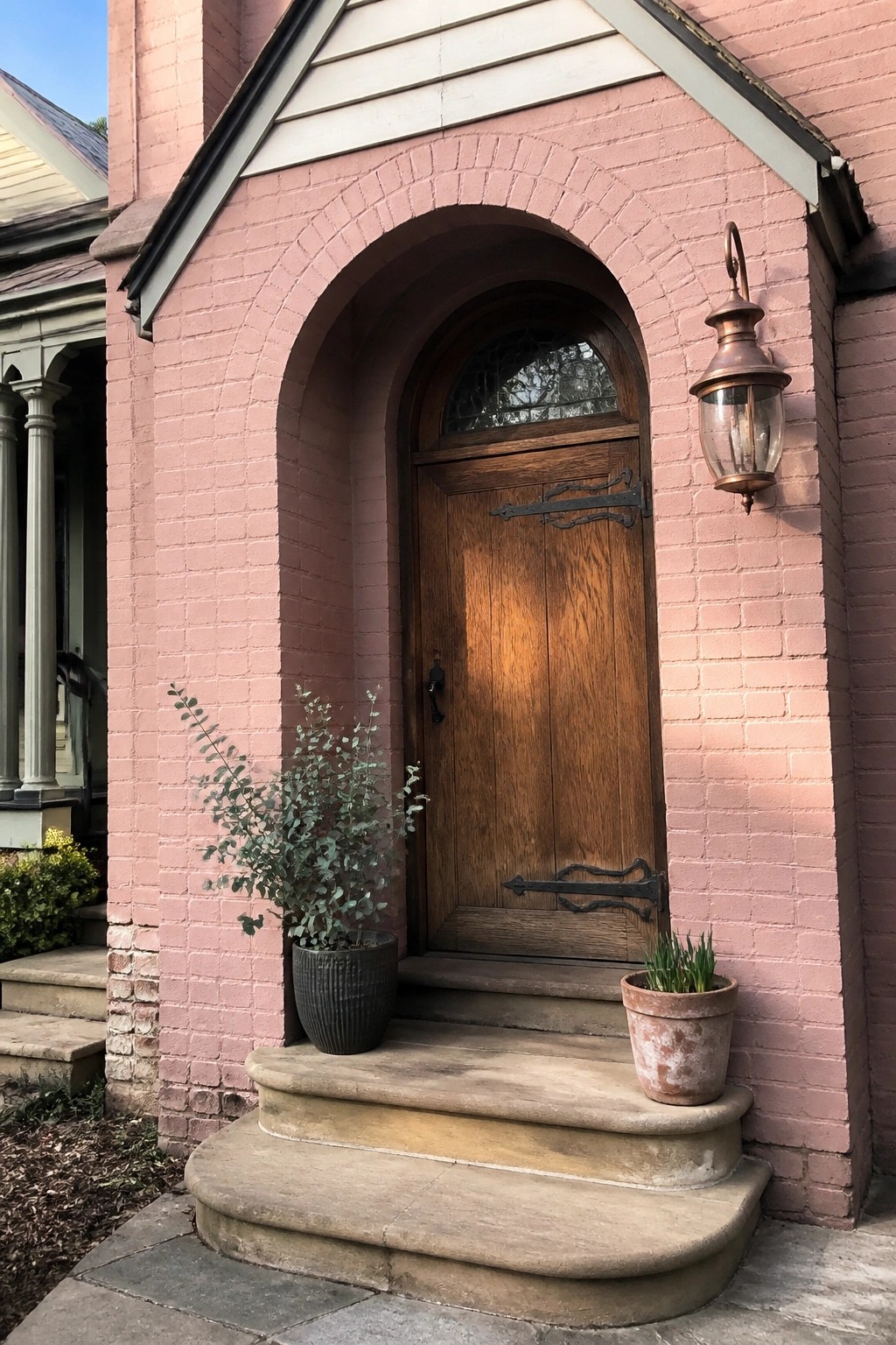 A pink brick house exterior featuring an arched brick entryway around a wooden front door, with concrete steps and two potted plants at the base.