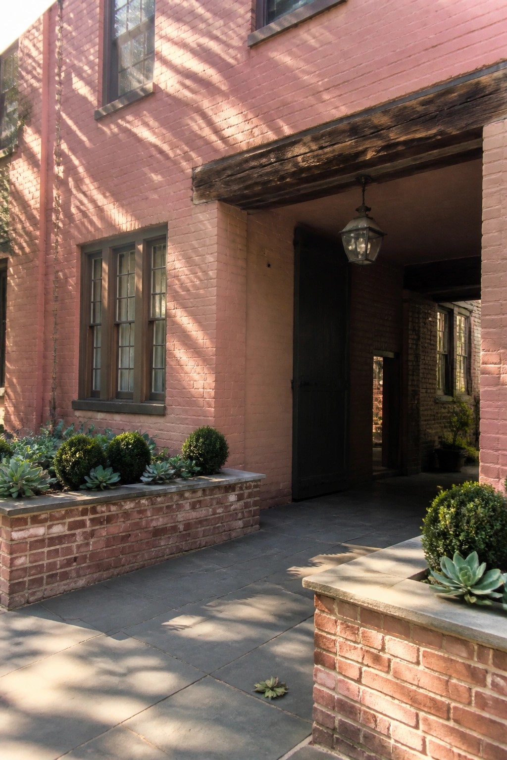 Pink brick house exterior with dark wood beam over entry, black door, and matching low brick planters holding green shrubs and succulents.
