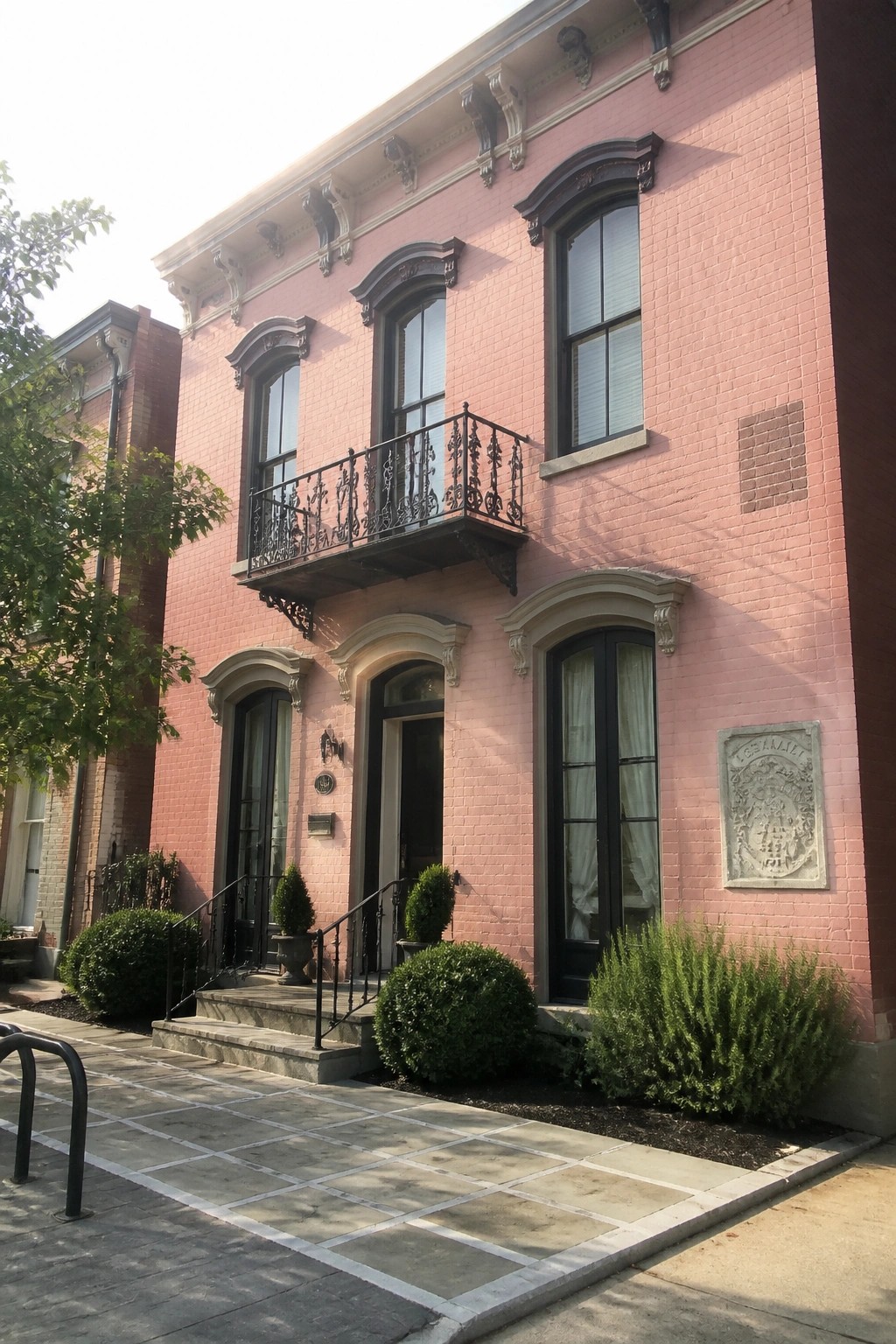 Pink brick townhouse with ornate black balcony, arched windows, and front shrubs