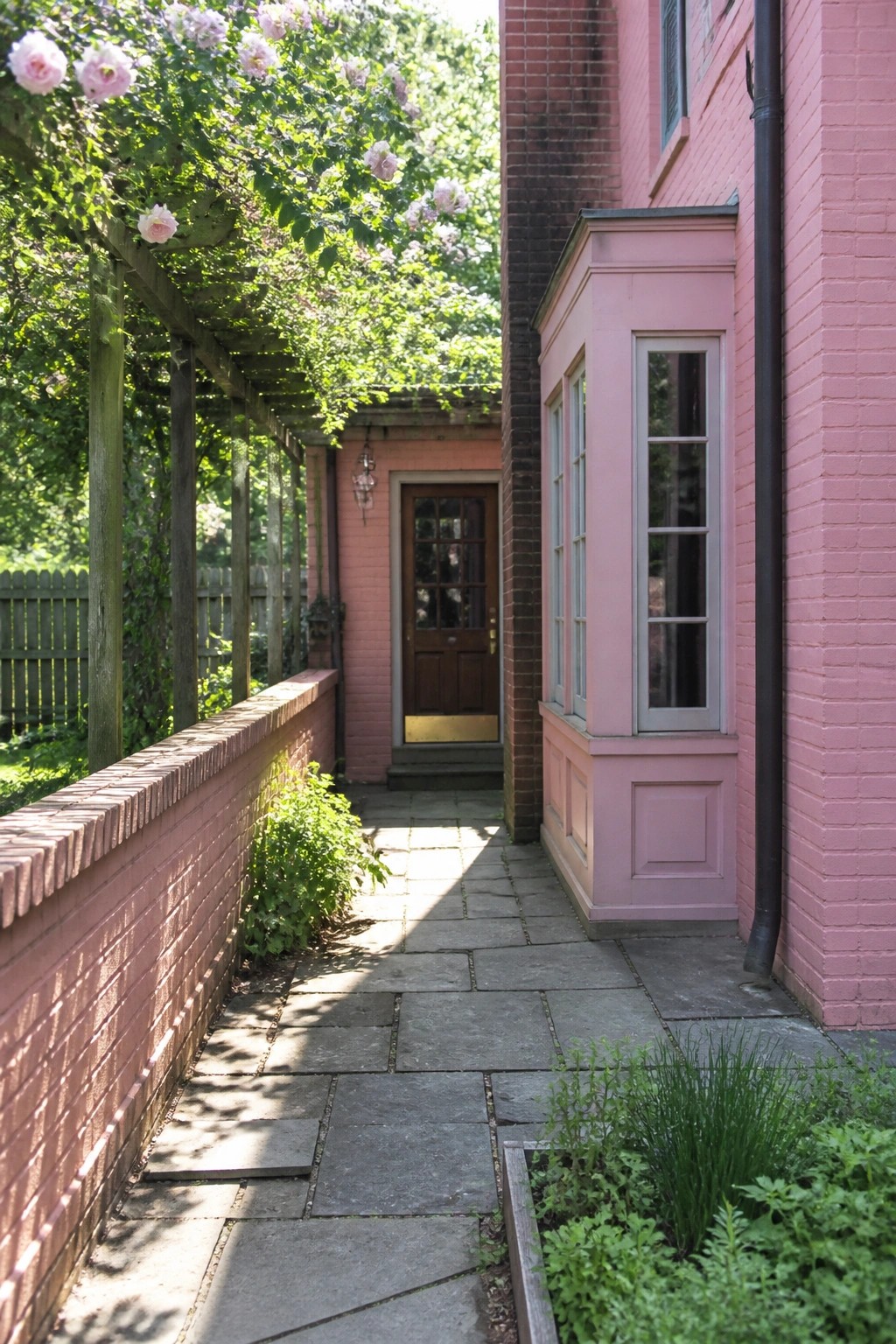 A narrow stone path runs alongside a pink brick house wall with a bay window and leads toward a brown wooden door.