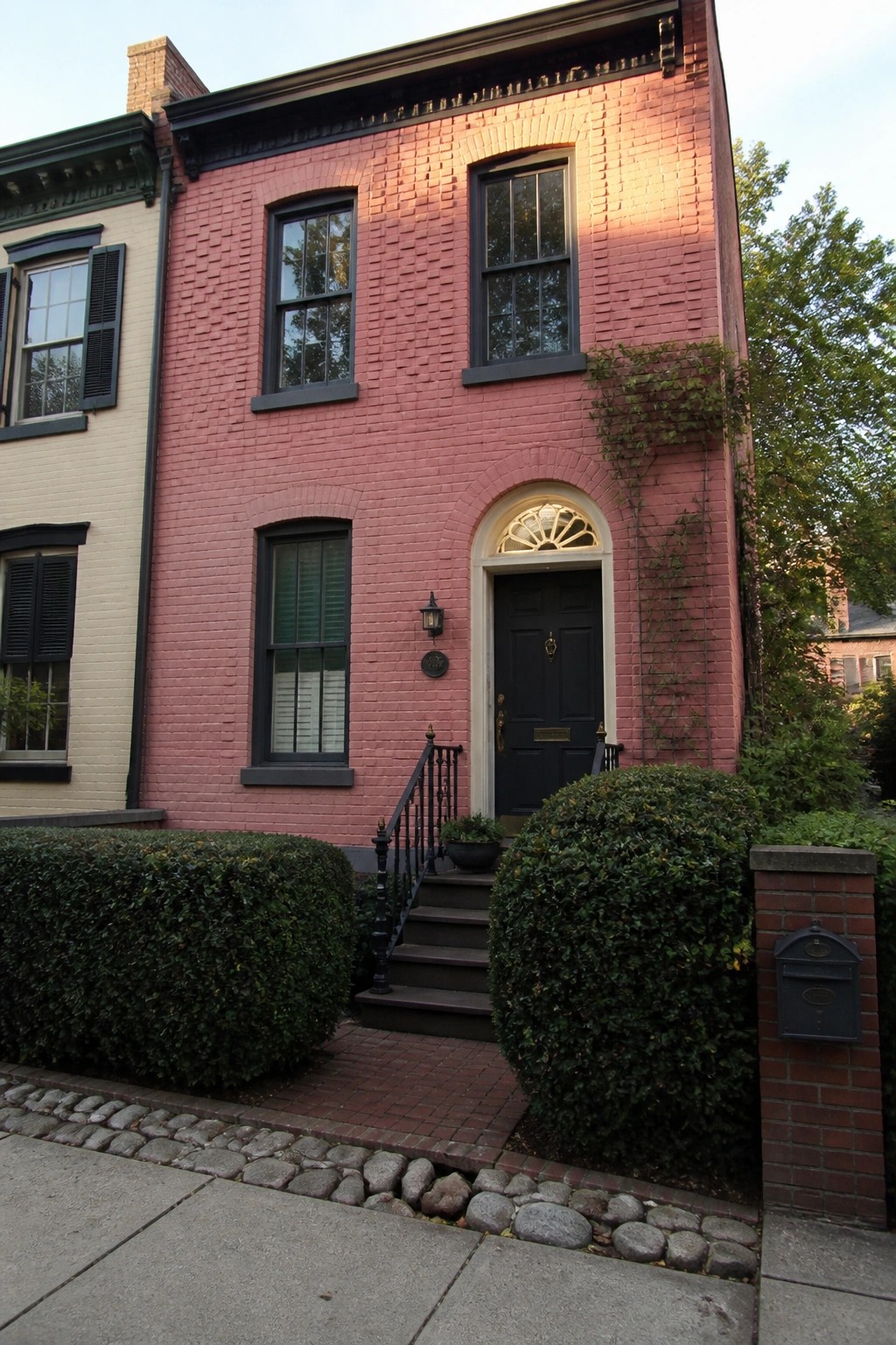 A two-story pink brick house with an arched entry, black door, and dark-trimmed windows, viewed from the sidewalk with shrubs in front.