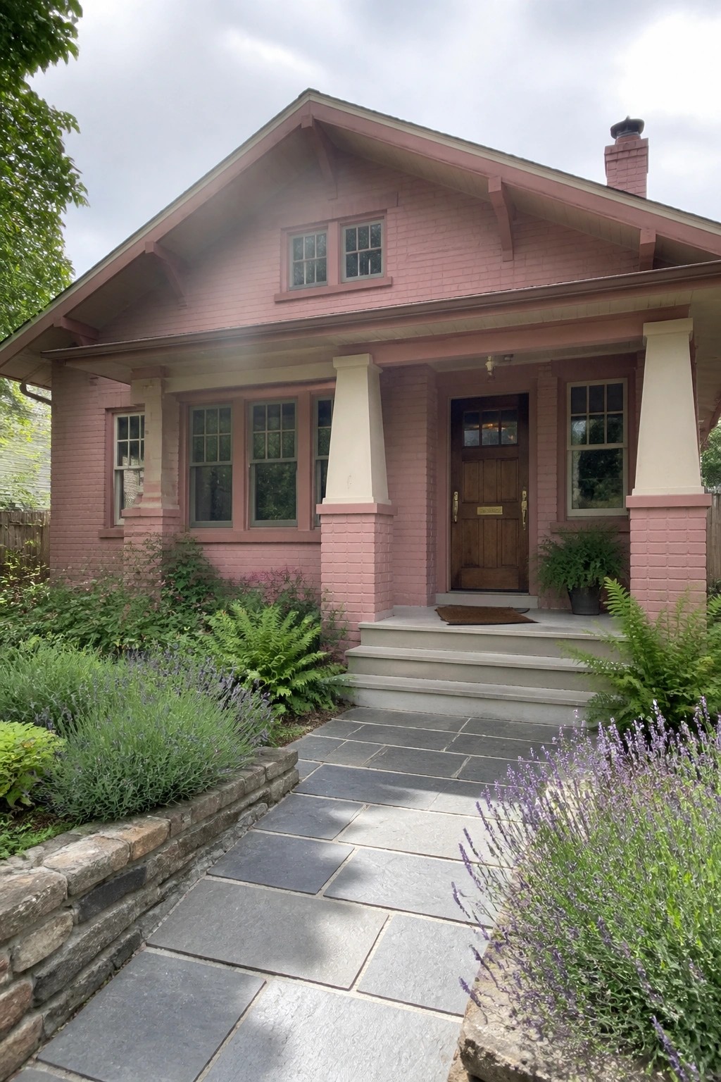 A single-story pink brick house with a gabled roof, front porch supported by white columns, wooden door, and stone walkway bordered by lavender and ferns.