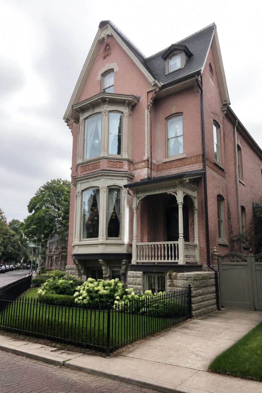 A two-and-a-half story pink brick house with a stone foundation, bay windows, a front porch, and a black iron fence in front.