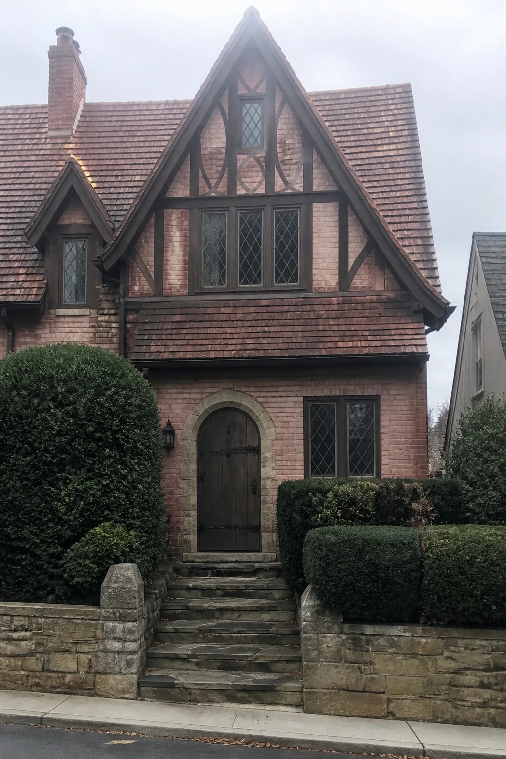 A two-story pink brick house with dark timber framing on the upper gable, an arched wooden front door, diamond-pane windows, and stone steps leading from the sidewalk.