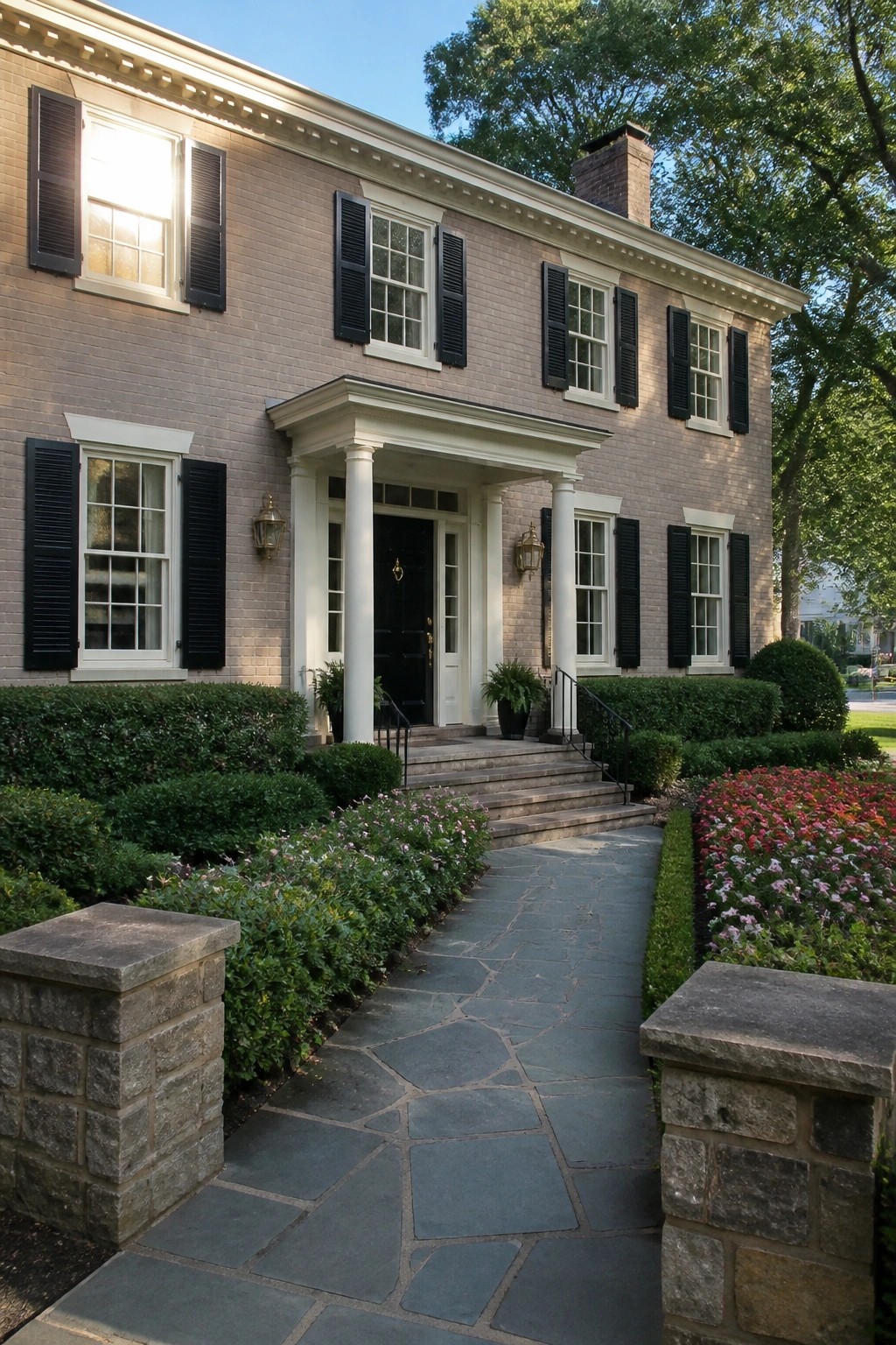 Two-story brick house with white columns, black shutters, stone walkway, and colorful flowers.