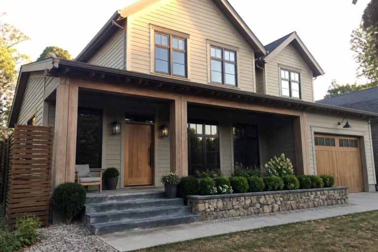 Front view of a two-story house with beige siding, a large wooden front door, exposed wooden porch beams, stone steps, and a wooden garage door.