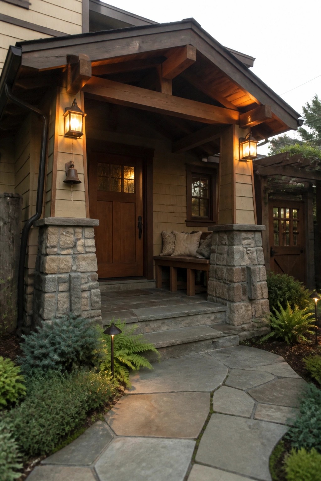 Front view of a beige house with a covered entry porch supported by stone pillars and dark wood beams, a wooden door, and a stone pathway with steps.