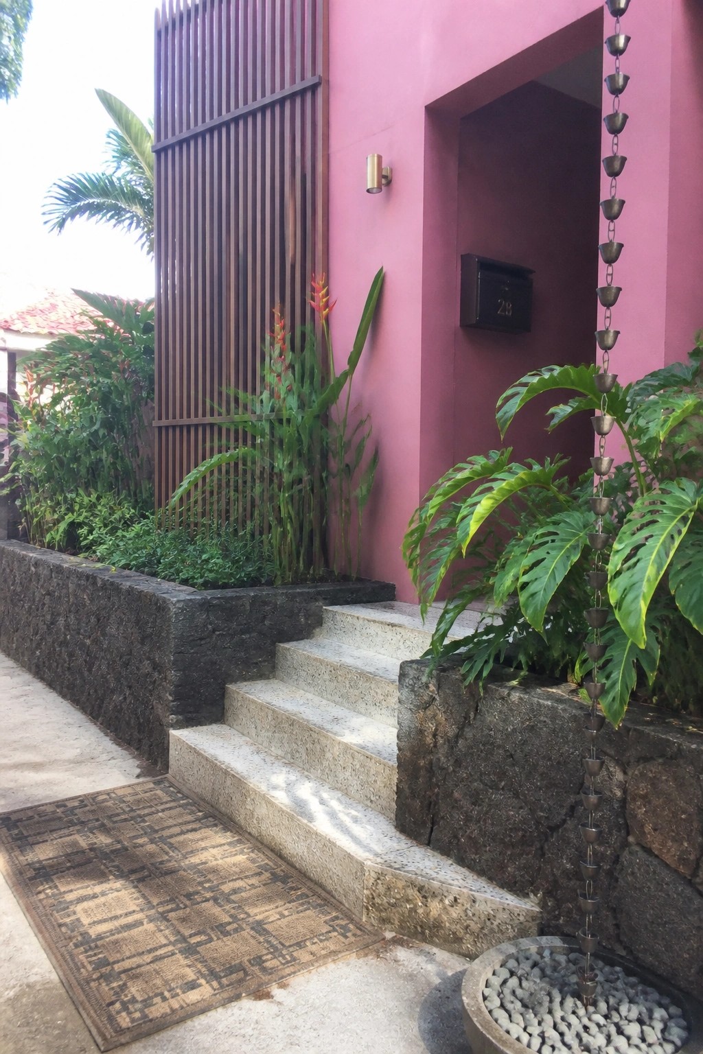 Pink house entrance with stone steps, doormat, tropical plants, and hanging rain chain.