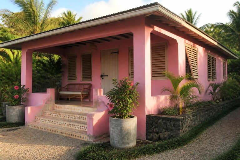 A single-story pink house with white trim, stone steps leading to a covered porch, and tropical plants along a gravel path.
