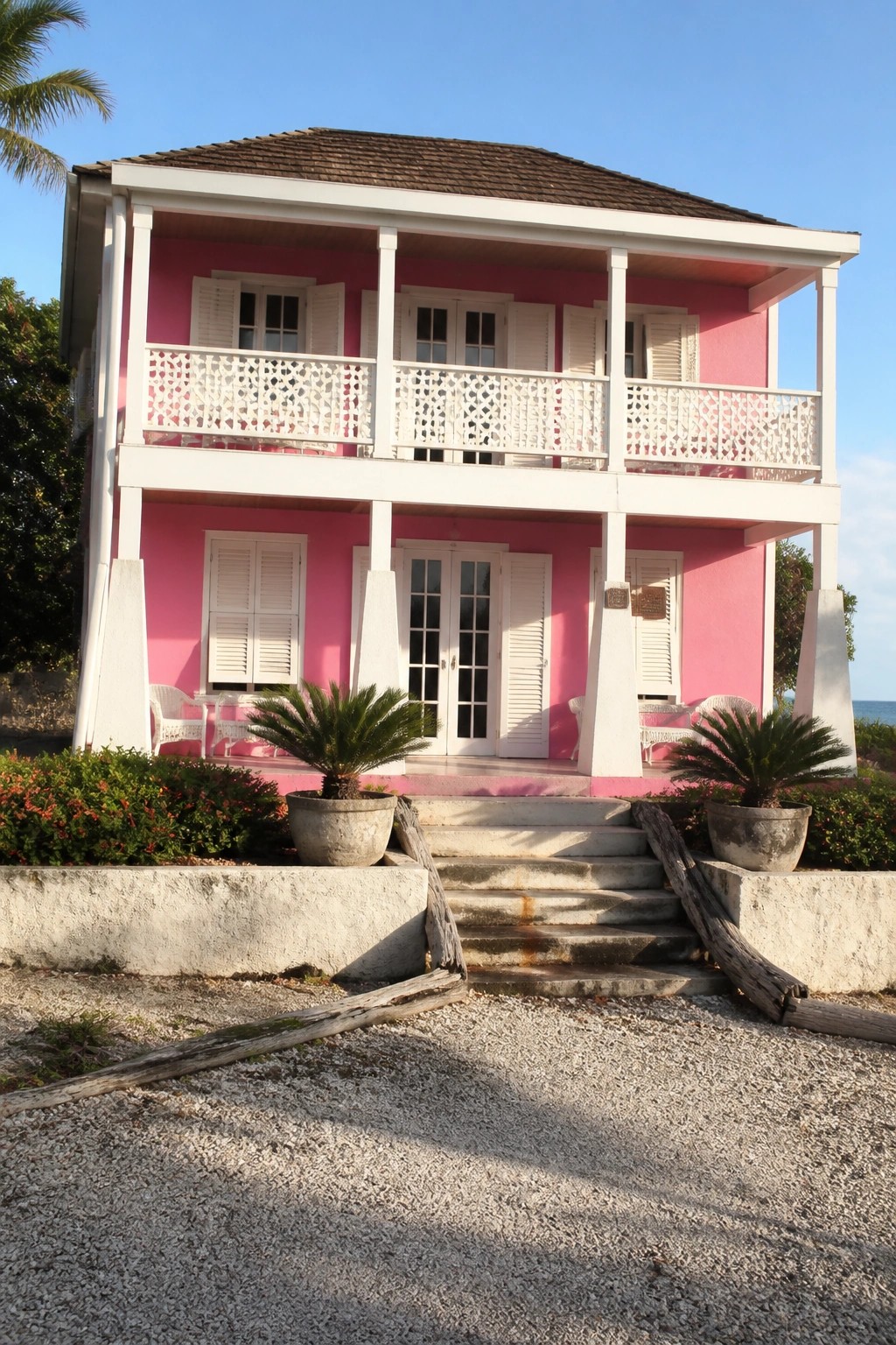 A two-story pink house with white trim, balconies, and a central staircase leading to double doors, set against a clear sky with ocean visible in the background.