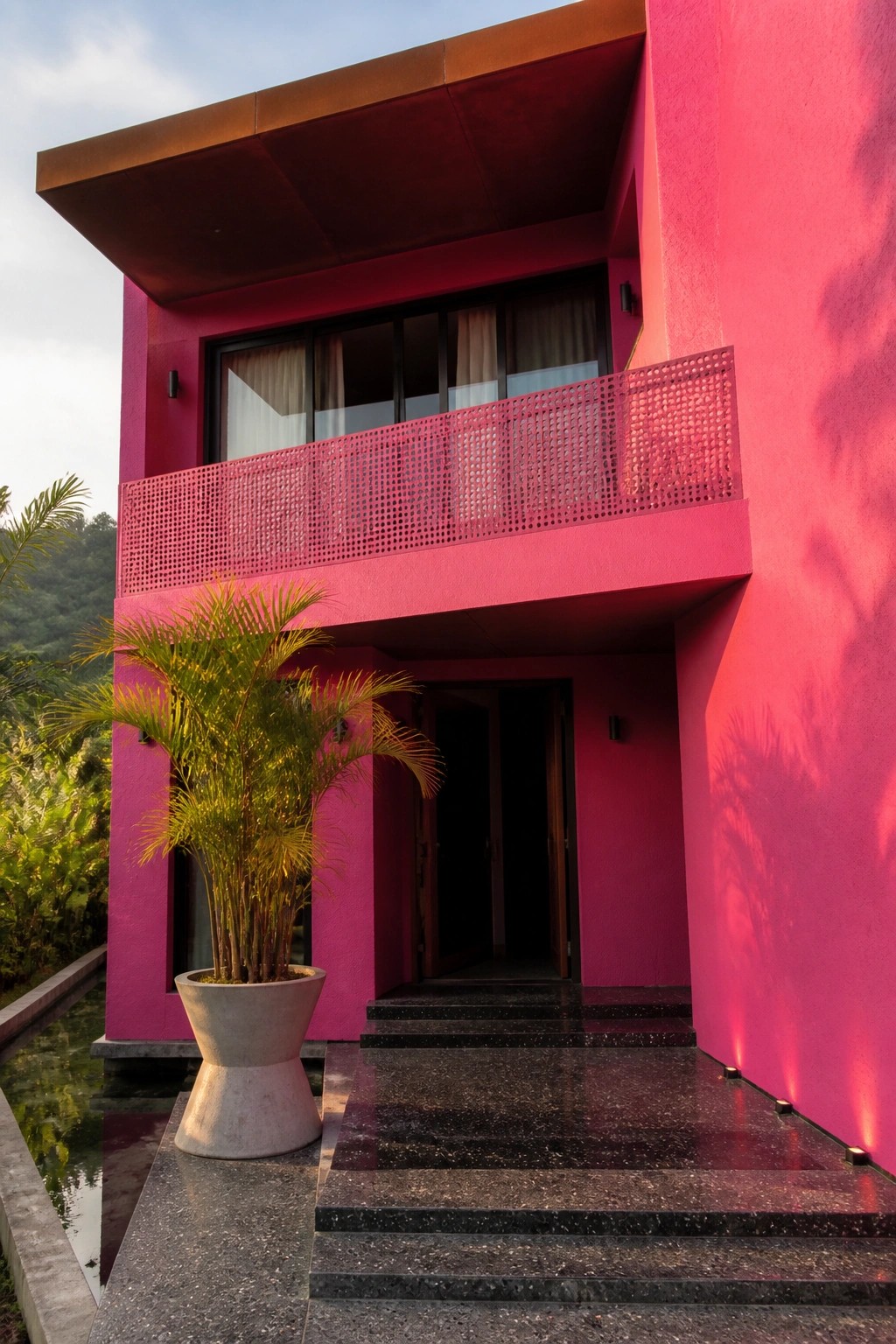 A modern two-story house with bright pink stucco walls, a perforated balcony railing, dark steps, an open doorway, and a potted palm near a small water feature.