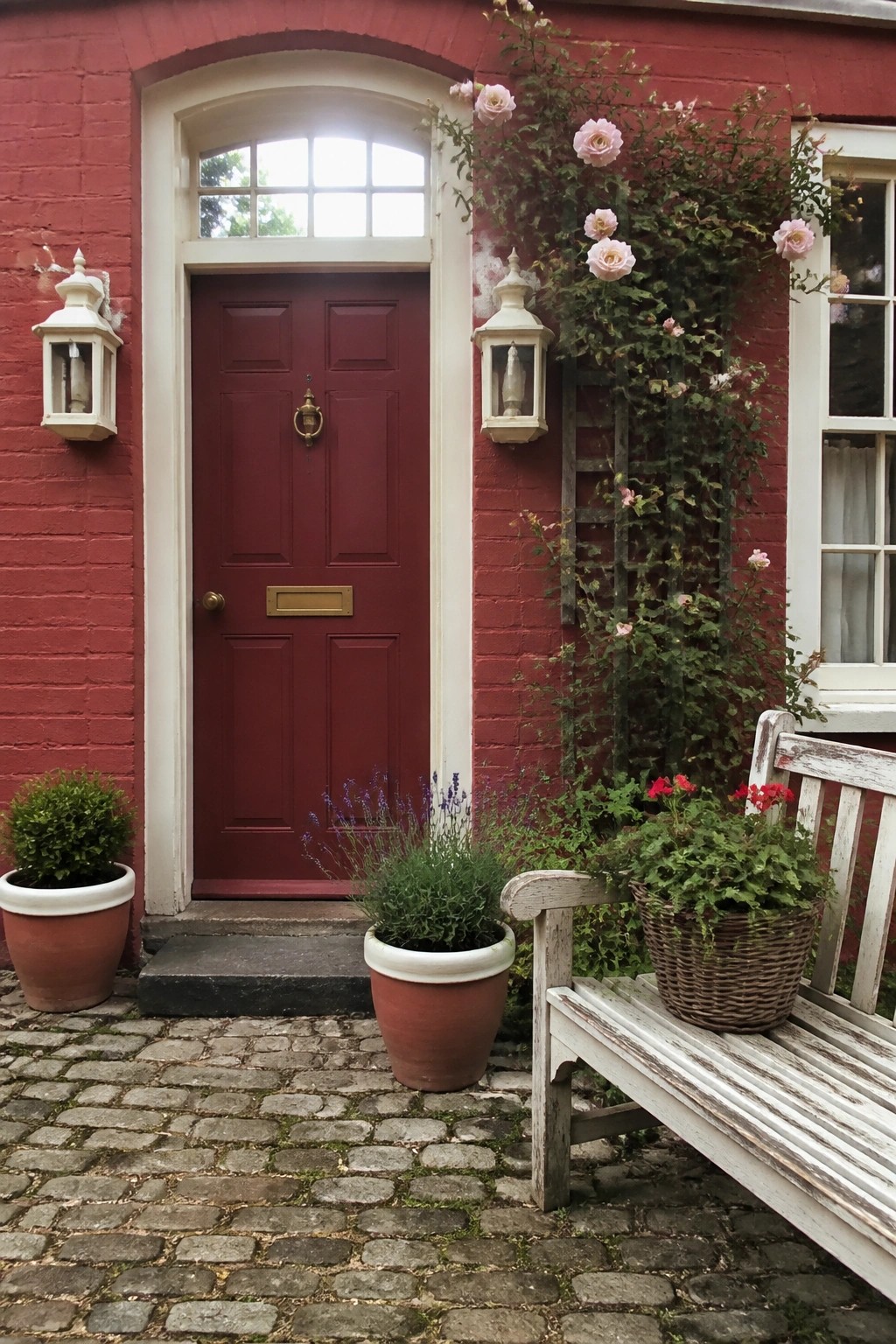 A red paneled door with cream trim on a brick house, white lanterns on both sides, and pink roses climbing a wooden trellis to the right of the door.