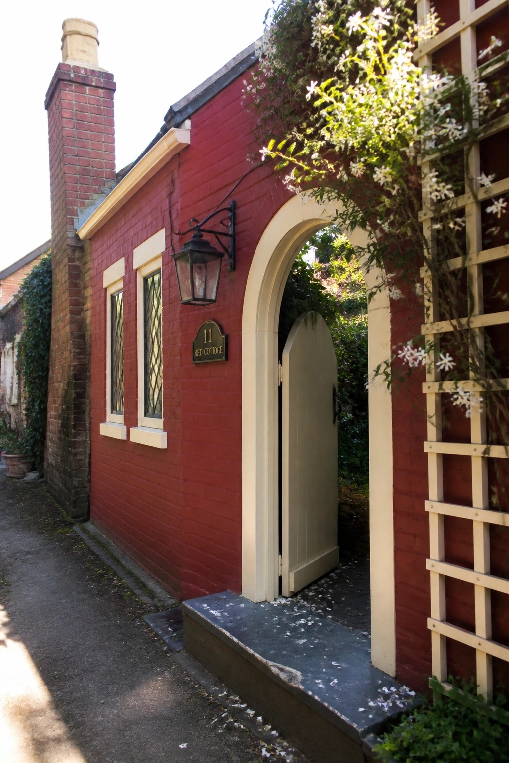 A red brick cottage with cream trim around an arched doorway, two small windows, and a light-colored arched door standing open.