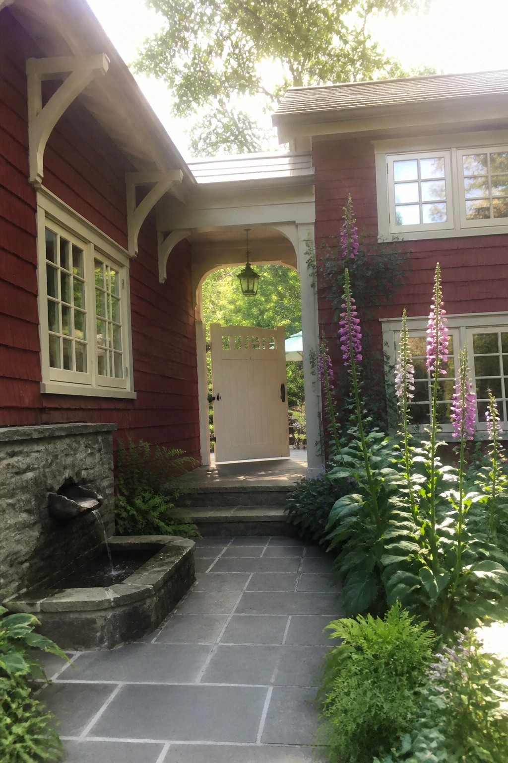 A red shingled cottage exterior with cream trim, an arched white entry gate, stone steps, and tall purple flowers along a paved path.