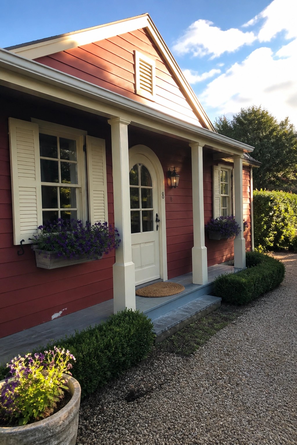 A red cottage exterior with cream trim on the windows, door surround, and porch columns, showing an arched front door and window boxes.