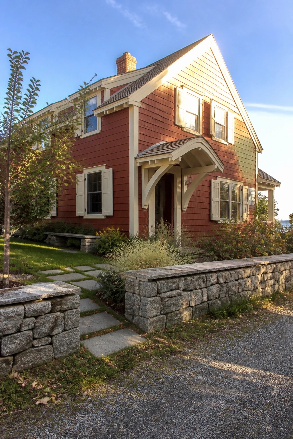 A two-story red shingled house with cream trim on the windows, door surround, and brackets, shown from the front with a stone wall and gravel path in the foreground.