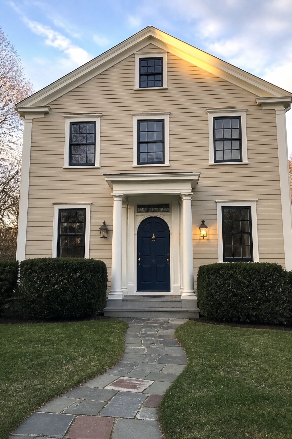 A two-story beige colonial house with white trim, a navy blue front door centered under a small portico, and a stone walkway leading to the steps.