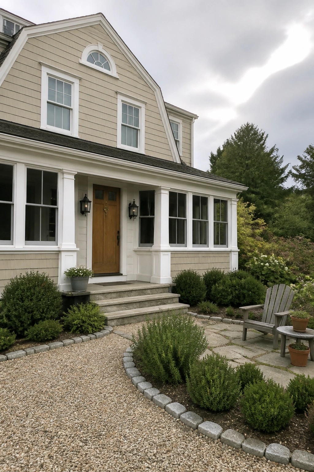 A beige colonial house with white trim, a wooden front door, stone steps, and a gravel path leading to a patio with shrubs and an Adirondack chair.