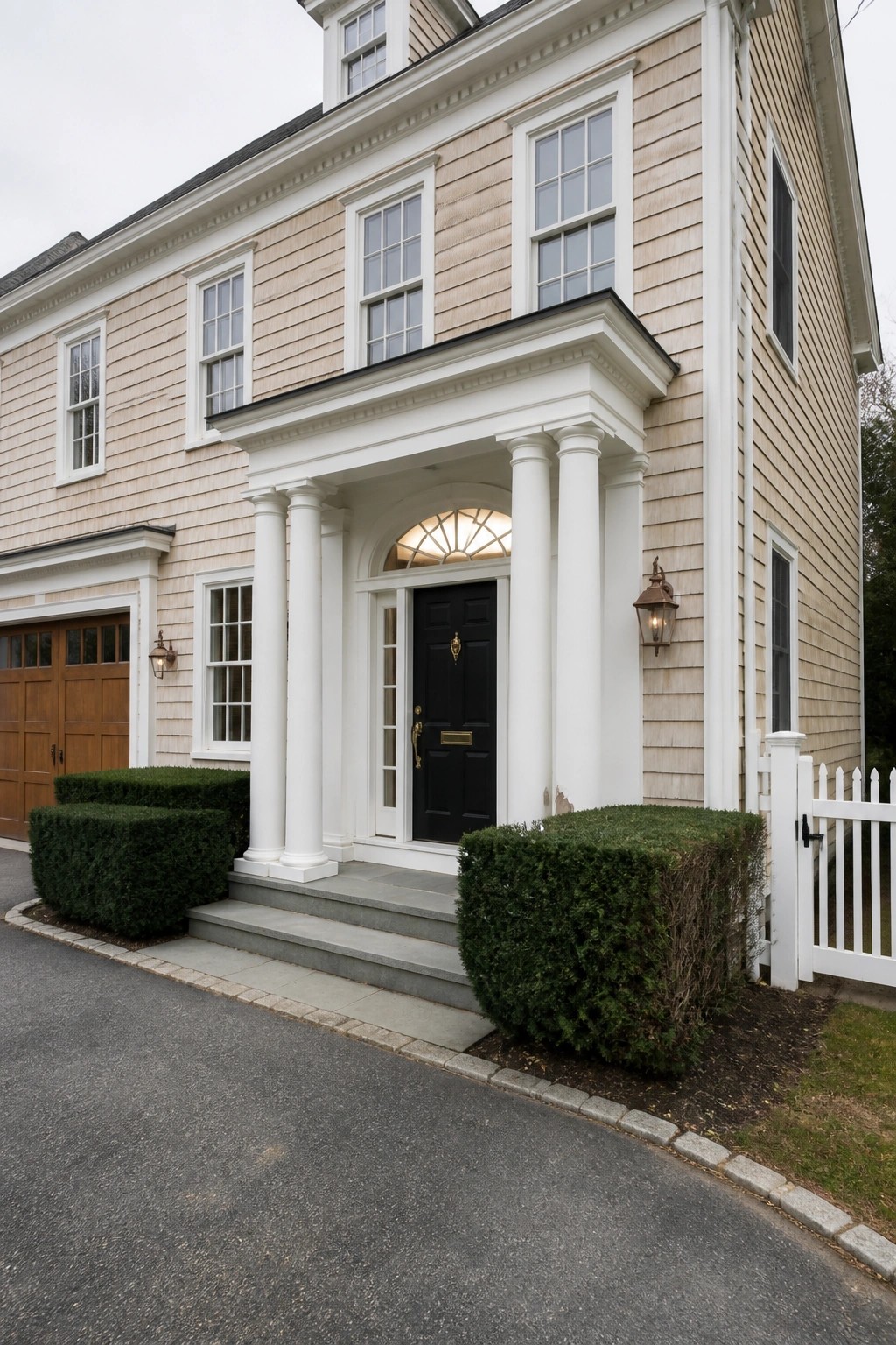 Beige shingled house with white columns, black door, and green bushes.