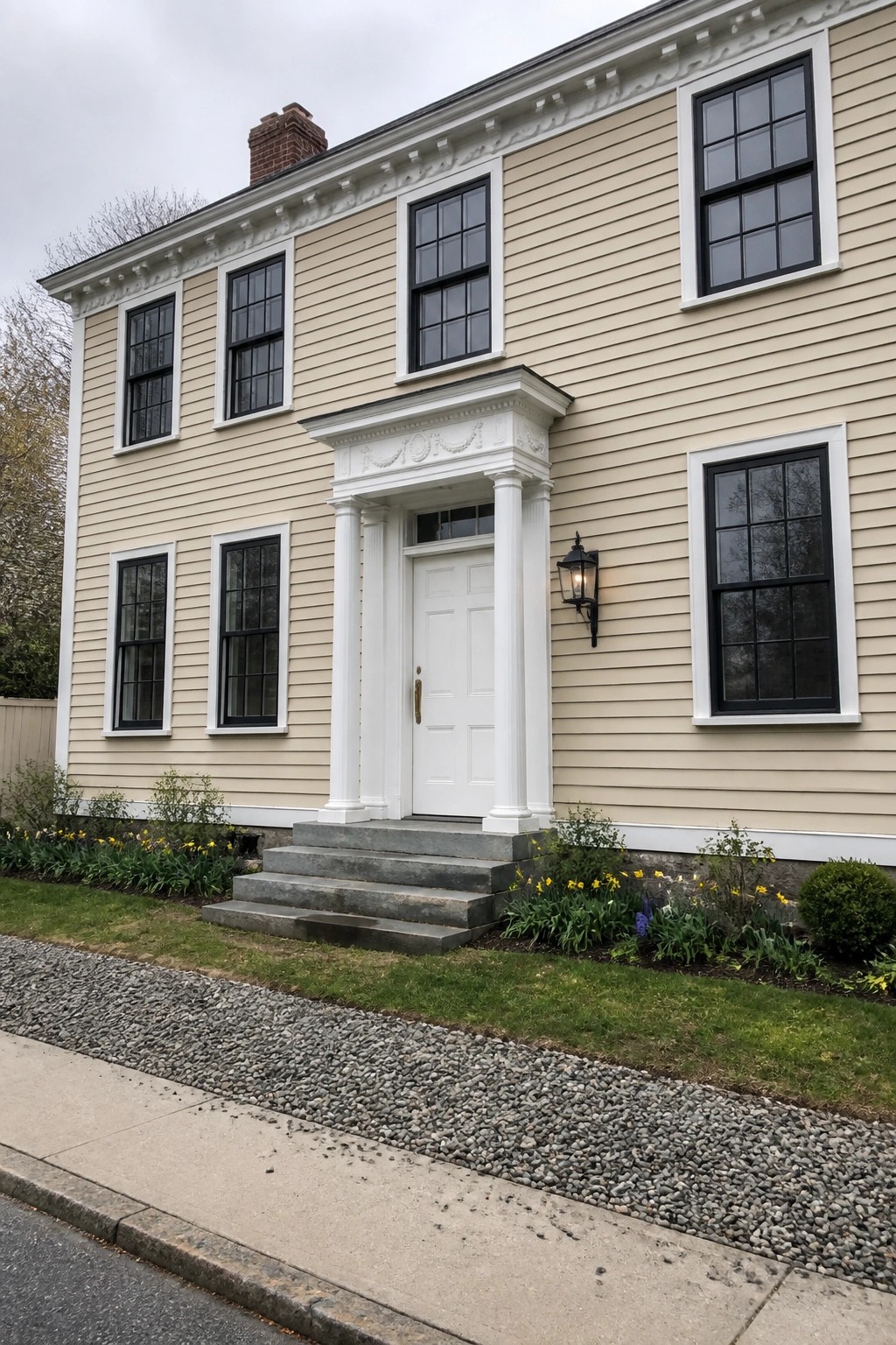 Two-story beige house with white columned door, black windows, and yellow flowers.
